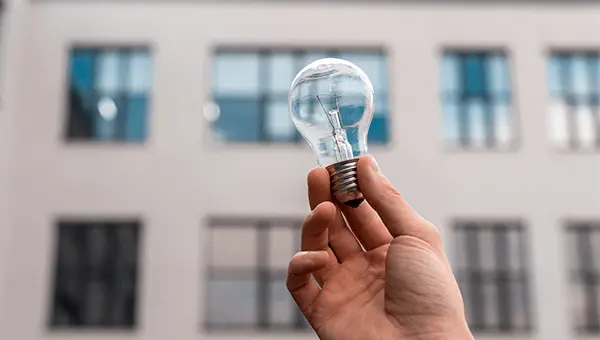 Żarówka trzymana w dłoni na tle elewacji budynku symbolizująca audyt energetyczny budynku.