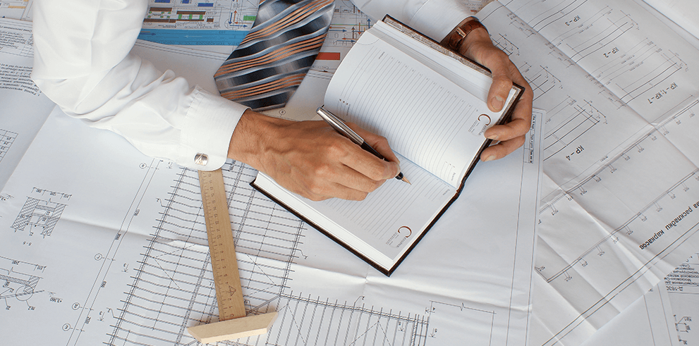 Osoba zapisująca notatki w notesie na biurku z rozłożonymi rysunkami technicznymi projektu budowlanego.