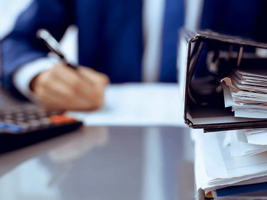 Segregator z dokumentami na biurku oraz osoba wypełniająca formularze. Kontrola dokumentacji na potrzeby ubezpieczenia IDI