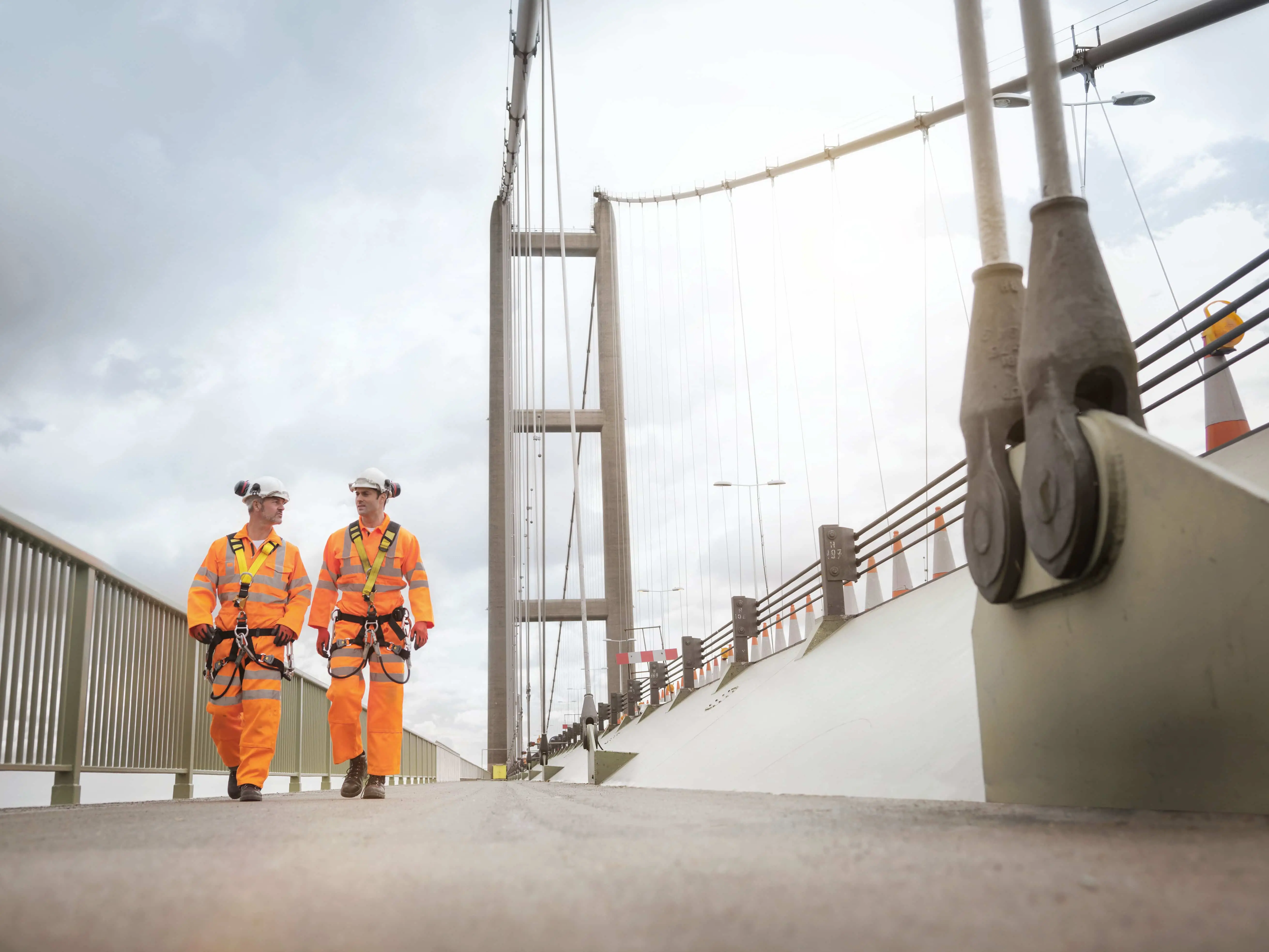 Kaksi työntekijää kävelee sillalla turvavarusteet päällä Kaksi työntekijää kävelee sillalla turvavarusteet päällä