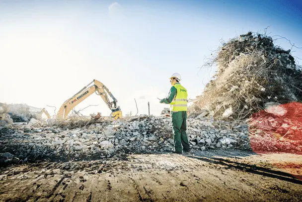 Person mit Schutzkleidung begutachtet Bauschutt, Bagger im Hintergrund