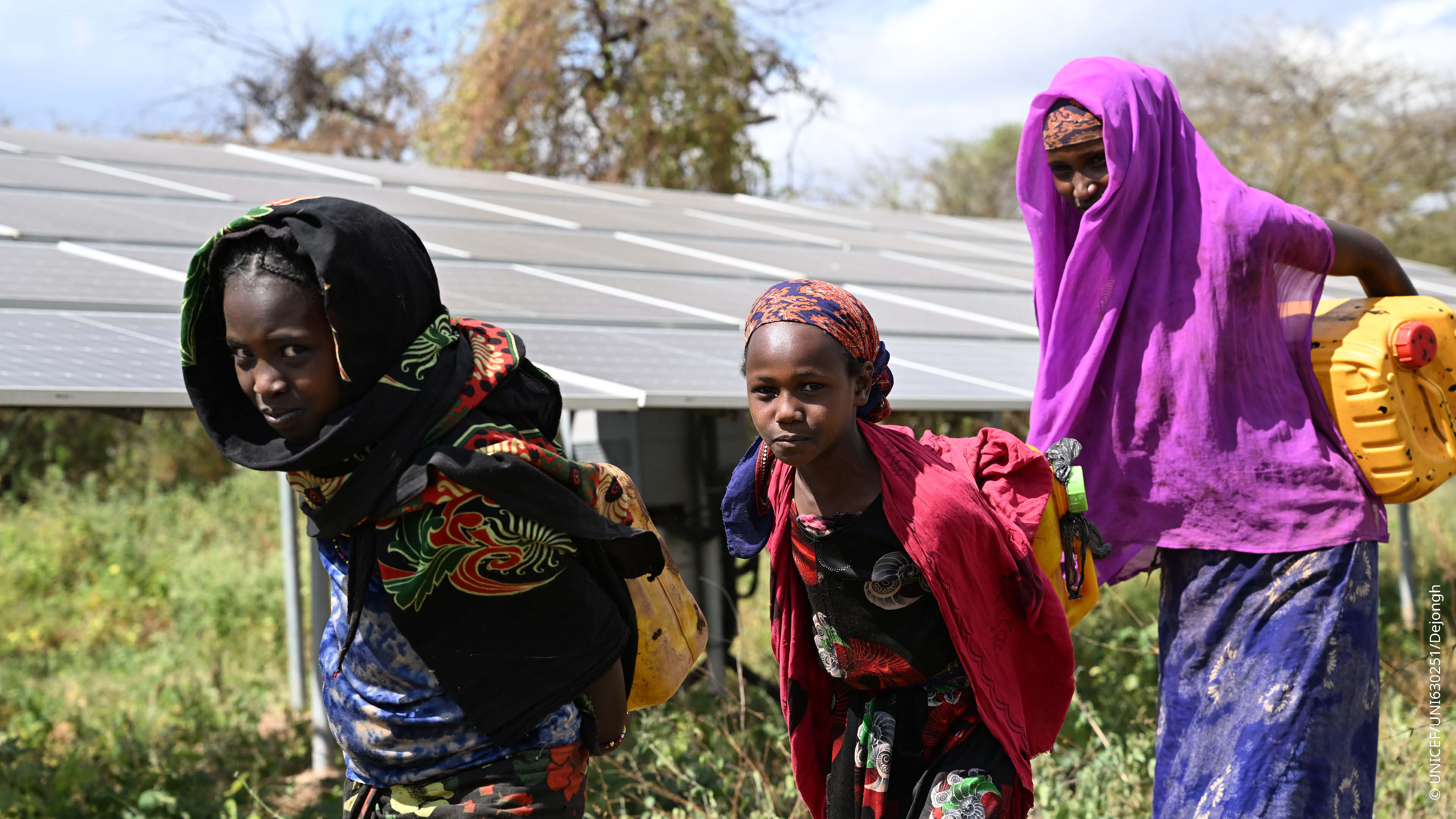 Three girls carry yellow water containers in front of an outdoor solar-powered water system. Three girls carry yellow water containers in front of an outdoor solar-powered water system.