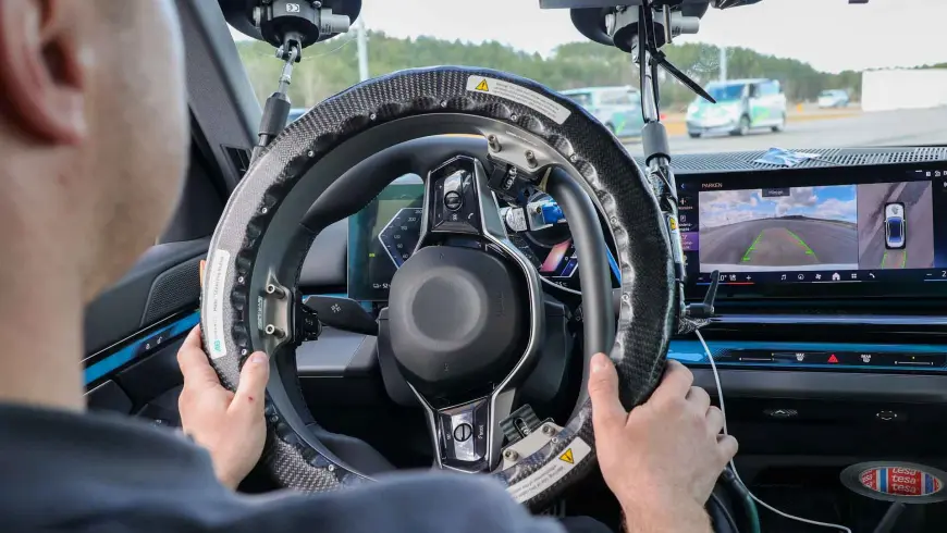  Test driver sits in the test vehicle behind a steering wheel equipped with driving robot technology, while monitors display driving data and camera images from the ADAS test runs at the Lausitzring.