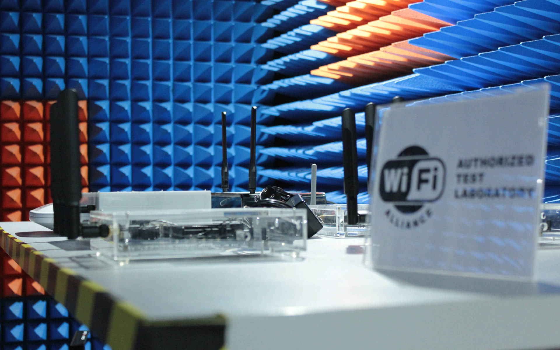 Test setup in a soundproof Wi-Fi chamber with antennas, devices, and a “Wi-Fi Authorized Test Laboratory” sign on a lab table. Test setup in a soundproof Wi-Fi chamber with antennas, devices, and a “Wi-Fi Authorized Test Laboratory” sign on a lab table.