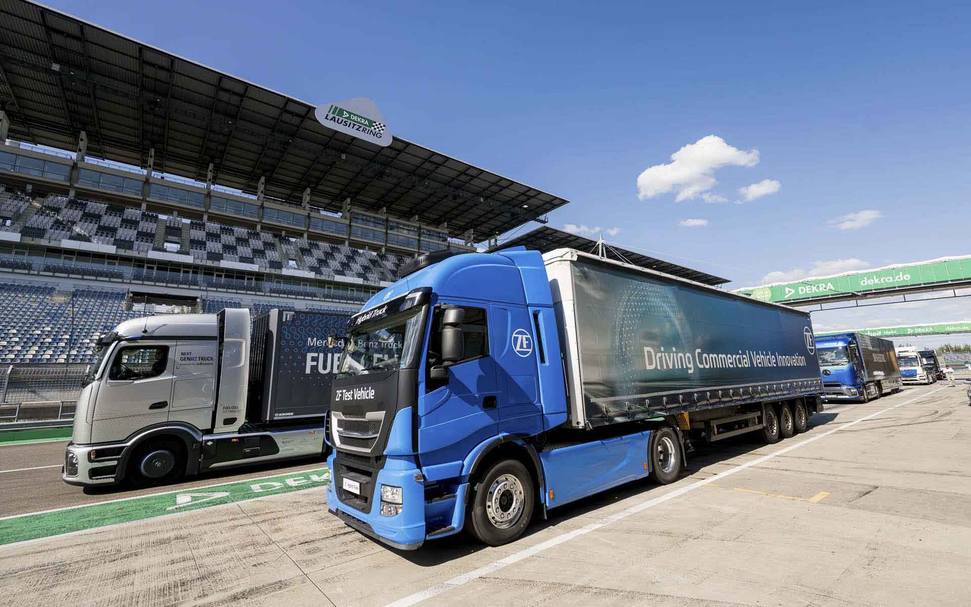 Mehrere batterieelektrische Lkw auf der Strecke des DEKRA Lausitzrings, dahinter die Haupttribüne. Mehrere batterieelektrische Lkw auf der Strecke des DEKRA Lausitzrings, dahinter die Haupttribüne.