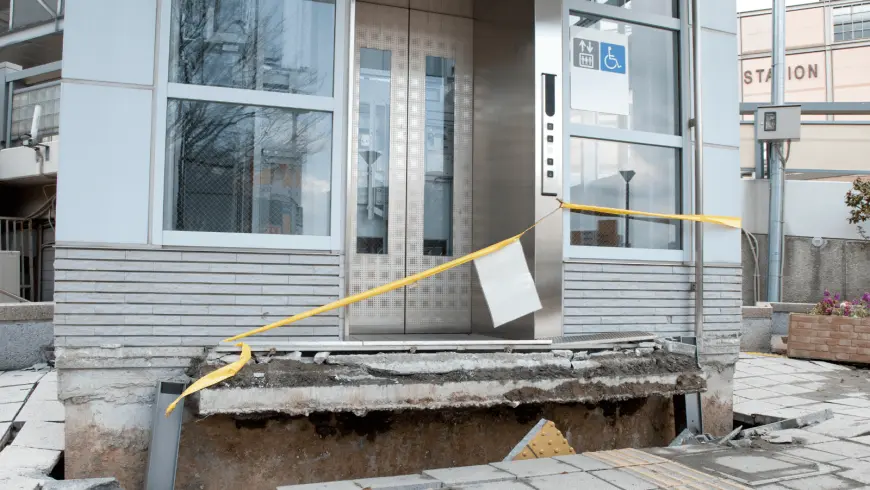 Exterior of a building with an elevator and a damaged concrete walkway, showing a large ground fissure likely caused by seismic activity. Yellow caution tape blocks off the area, and a station sign is visible in the background.