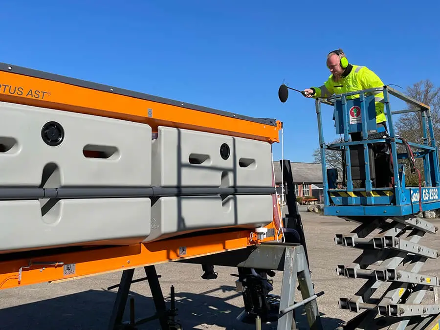 A man standing on a pallet jack conducting acoustic measurements using a microphone.