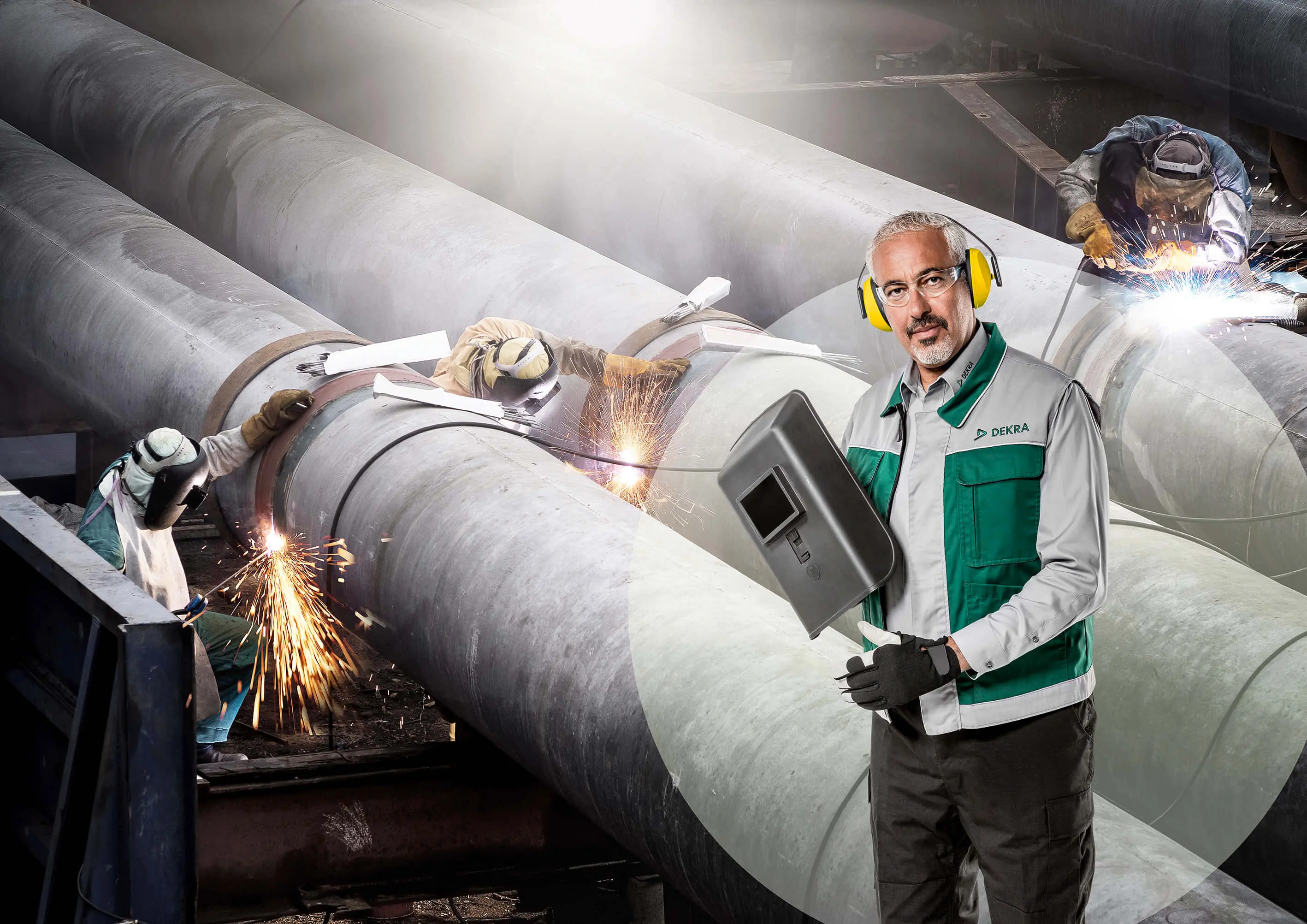DEKRA-tarkastaja valvoo hitsaajia, jotka työskentelevät suurten teollisuusputkien parissa, varmistaen vaatimustenmukaisuuden.
