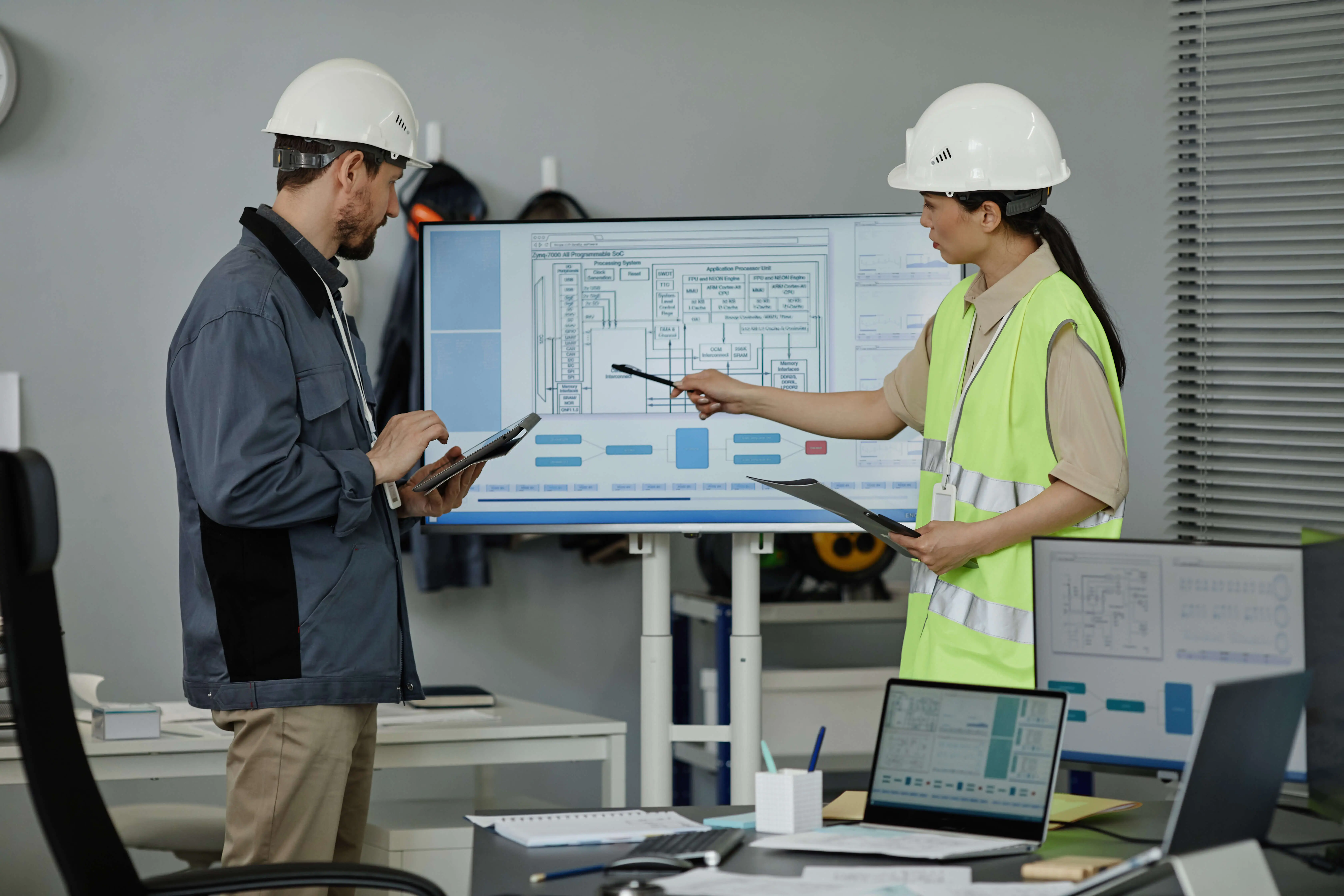 Two persons wearing safety helmets pointing on a screen where a plan is showen