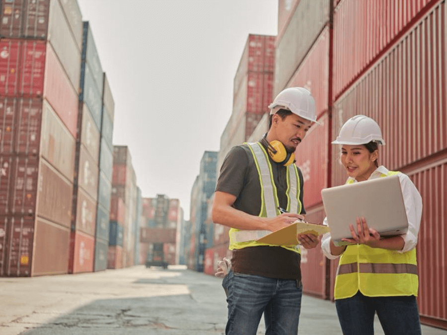 two people wearing protective helmets look at plans at a storage yard with containers in the background two people wearing protective helmets look at plans at a storage yard with containers in the background