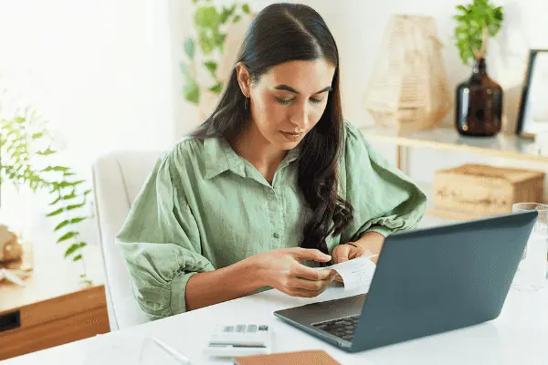 A woman is sitting at a desk, in front of a computer. She is looking at the bills she is holding.