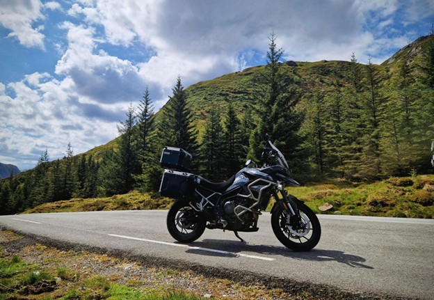 Odstavená motorka na ceste pod modrou oblohou, zelené kopce a stromy v pozadí