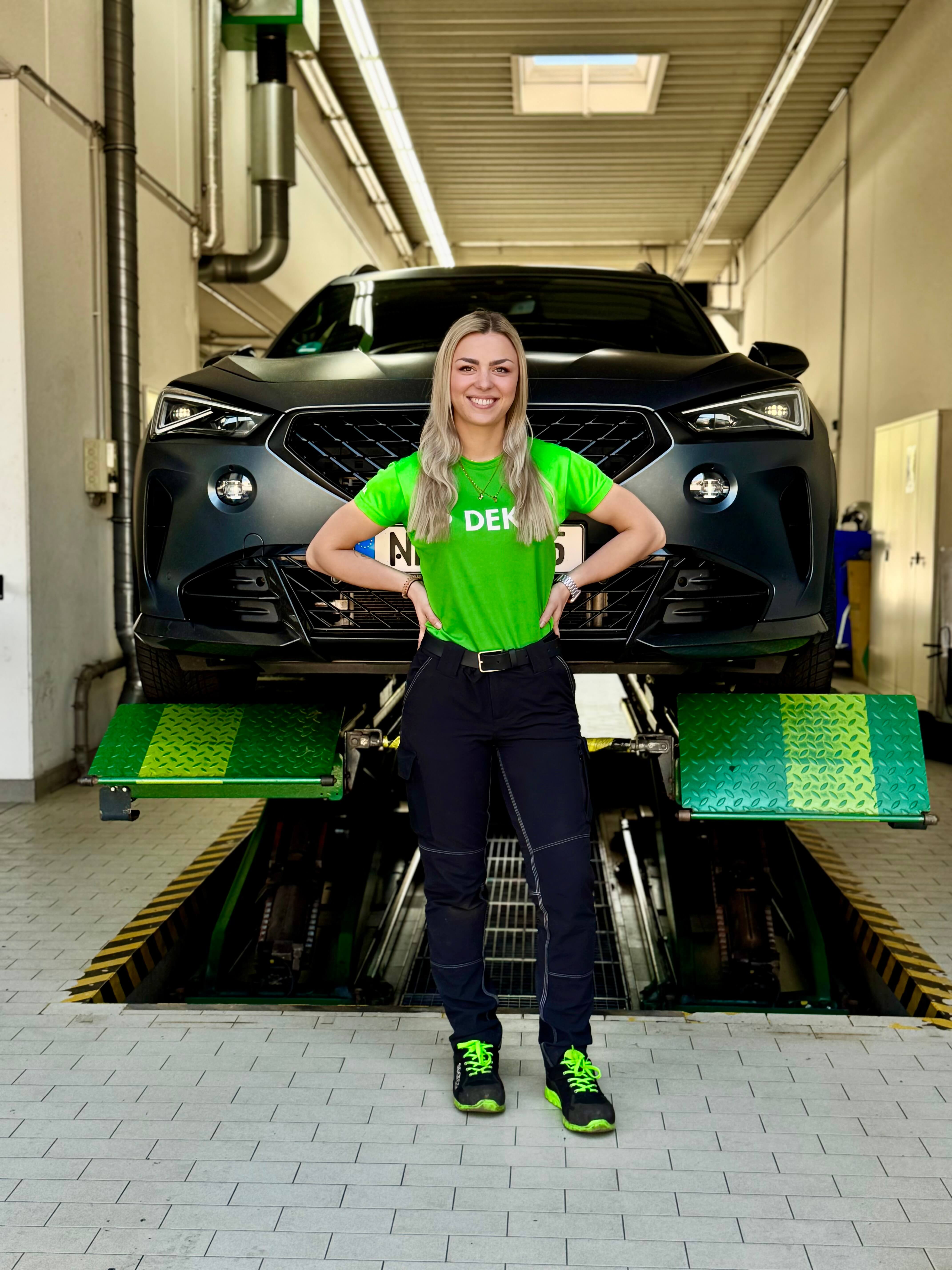 Michelle, Kfz Prüfingenieurin bei DEKRA, steht in Arbeitskleidung in der Prüfhalle vor Auto auf Hebebühne Michelle, Kfz Prüfingenieurin bei DEKRA, steht in Arbeitskleidung in der Prüfhalle vor Auto auf Hebebühne