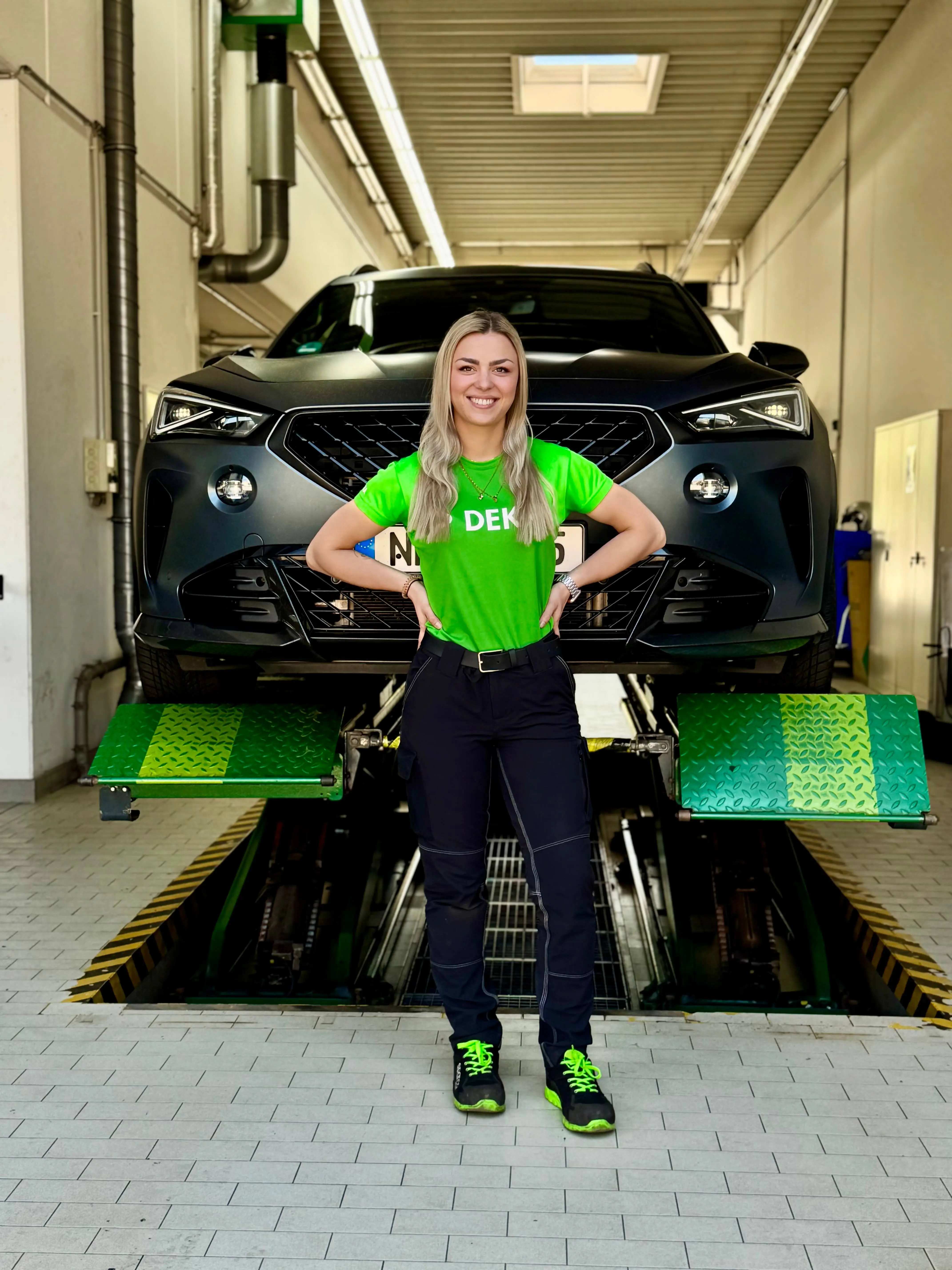 Michelle, Kfz Prüfingenieurin bei DEKRA, steht in Arbeitskleidung in der Prüfhalle vor Auto auf Hebebühne