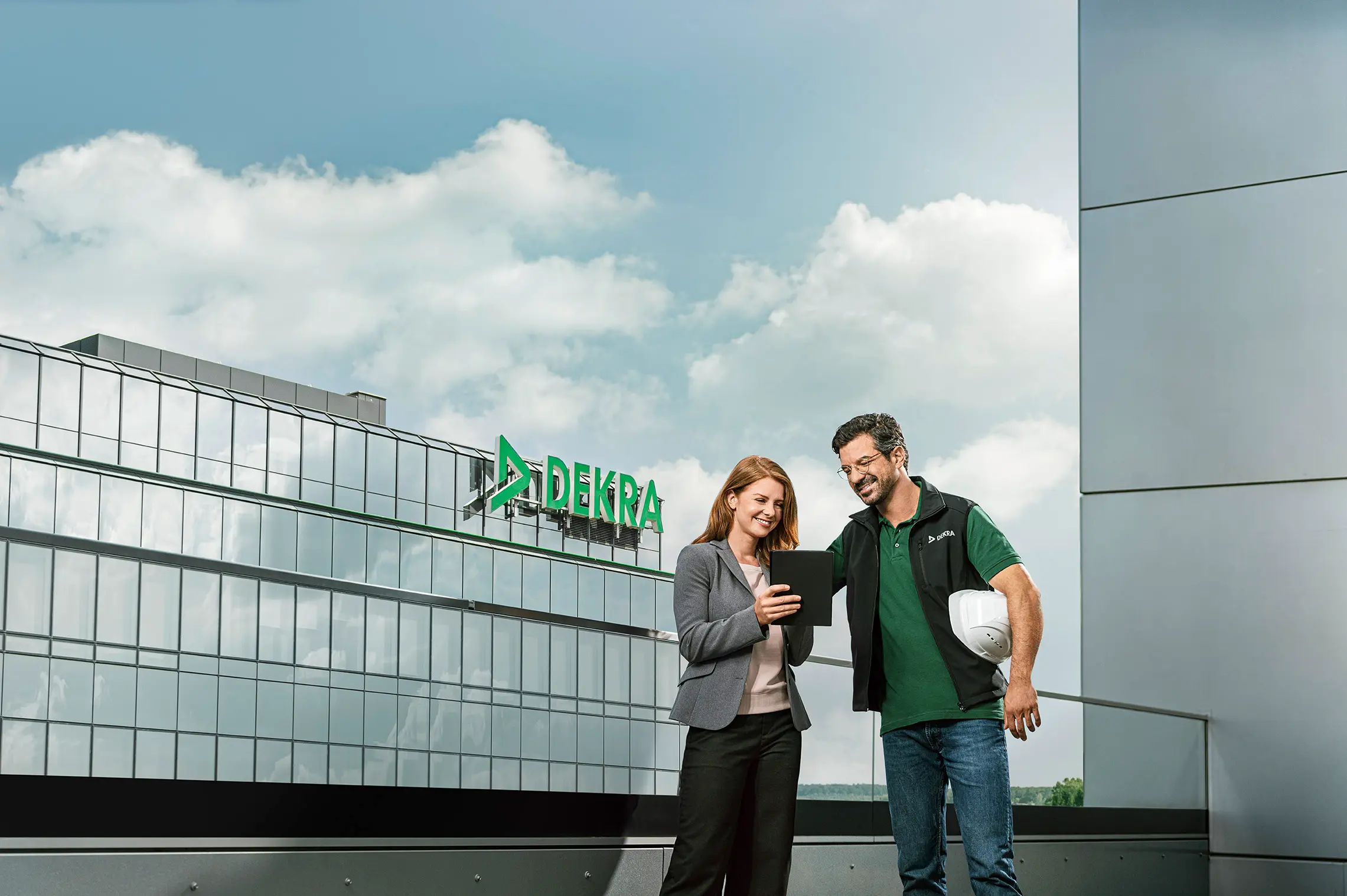 Mann in DEKRA Arbeitskleidung und Frau im Business-Look schauen lächelnd auf ein Tablet vor einem DEKRA Gebäude