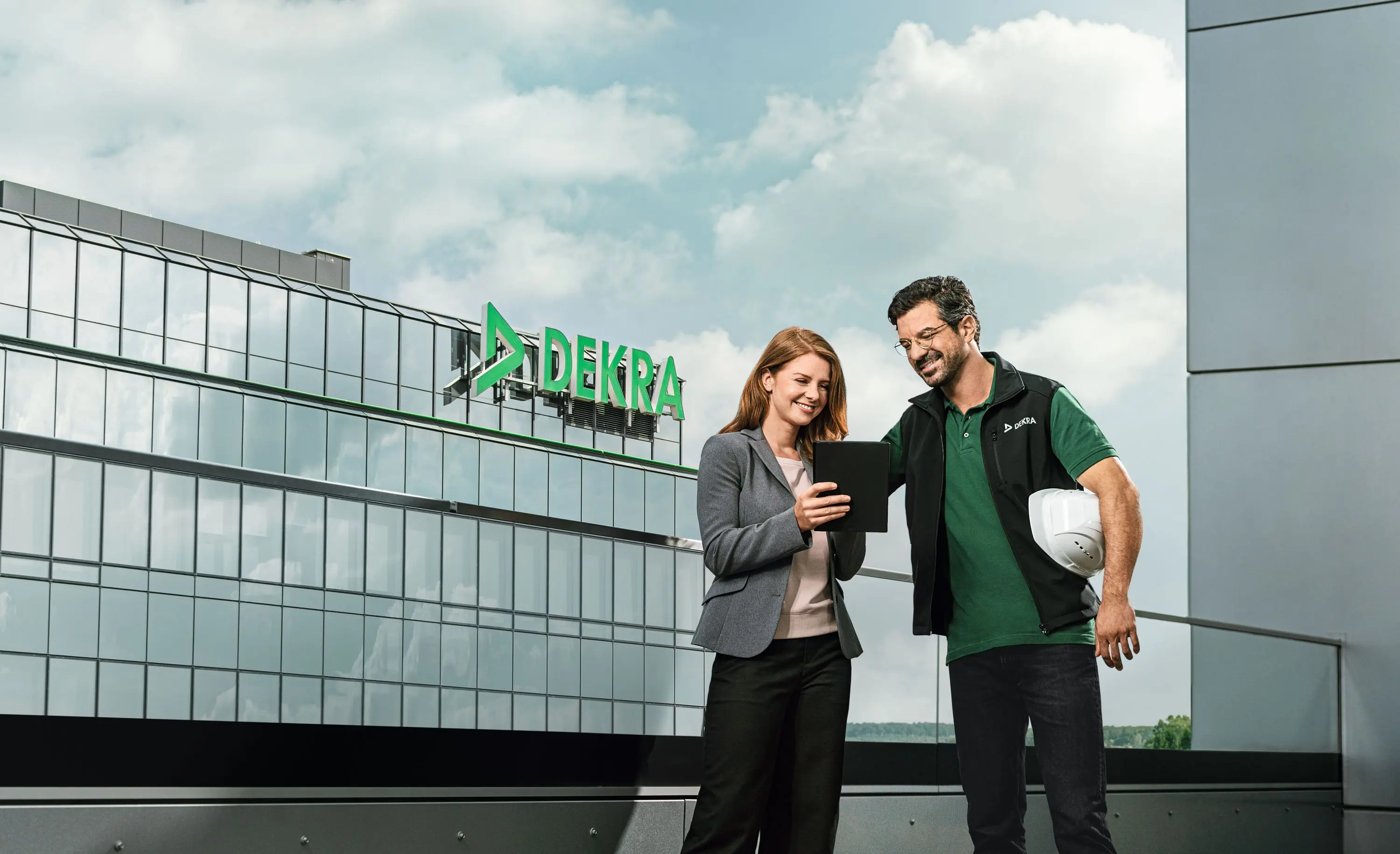 DEKRA Mitarbeiter und Kollegin mit Tablet vor Firmengebäude, lächelnd