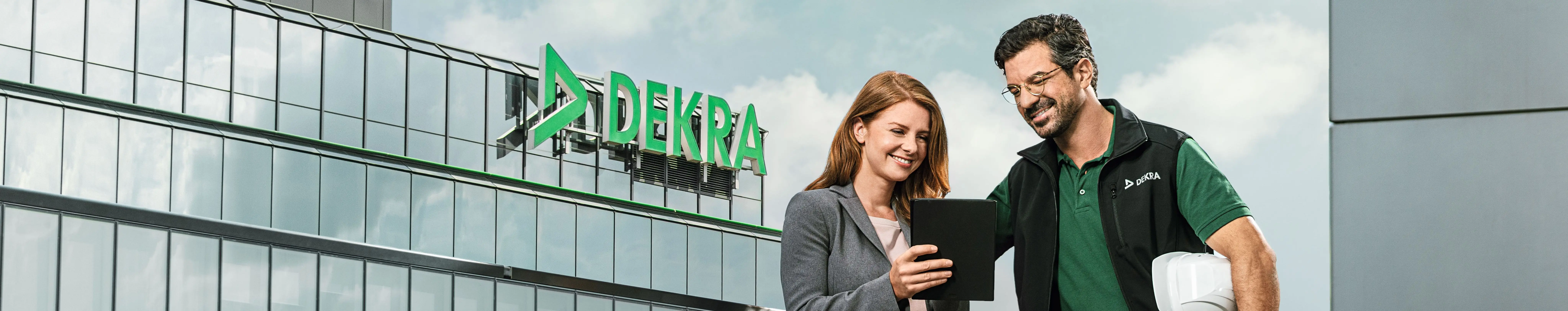 Mann in DEKRA Arbeitskleidung und Frau im Business-Look schauen lächelnd auf ein Tablet vor einem DEKRA Gebäude