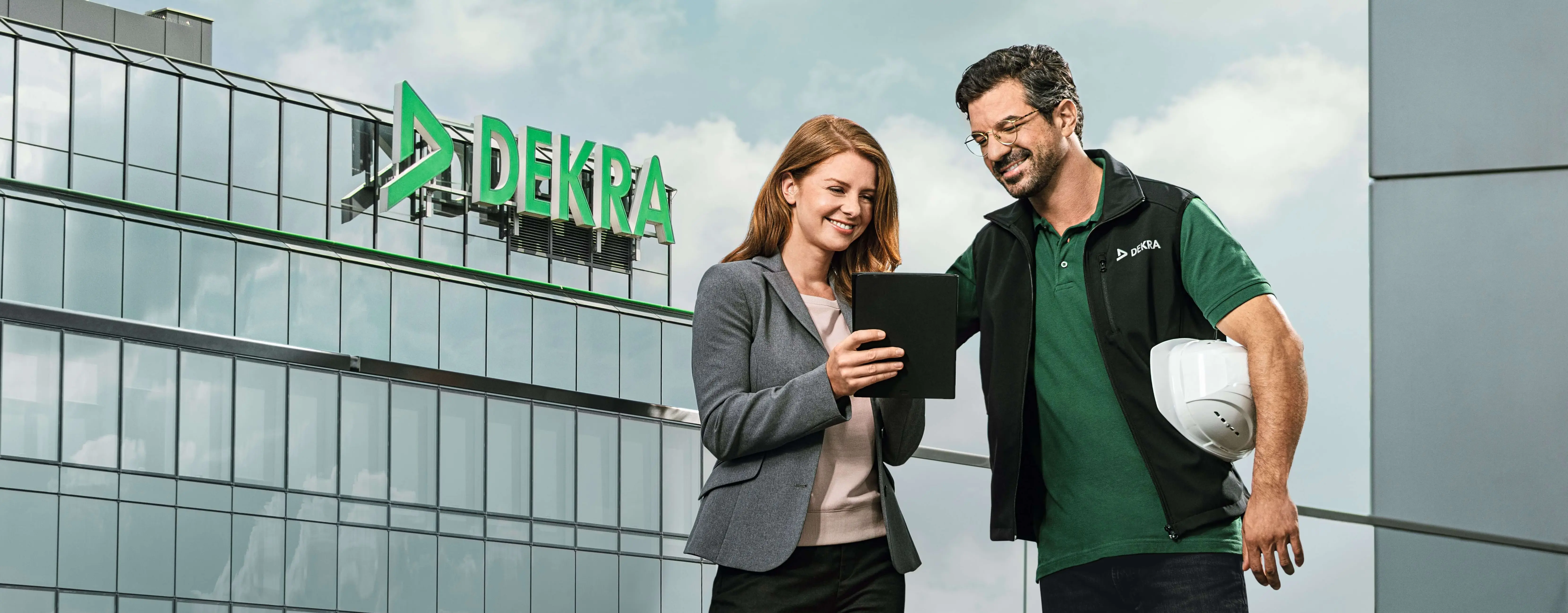 DEKRA Mitarbeiter und Kollegin mit Tablet vor Firmengebäude, lächelnd