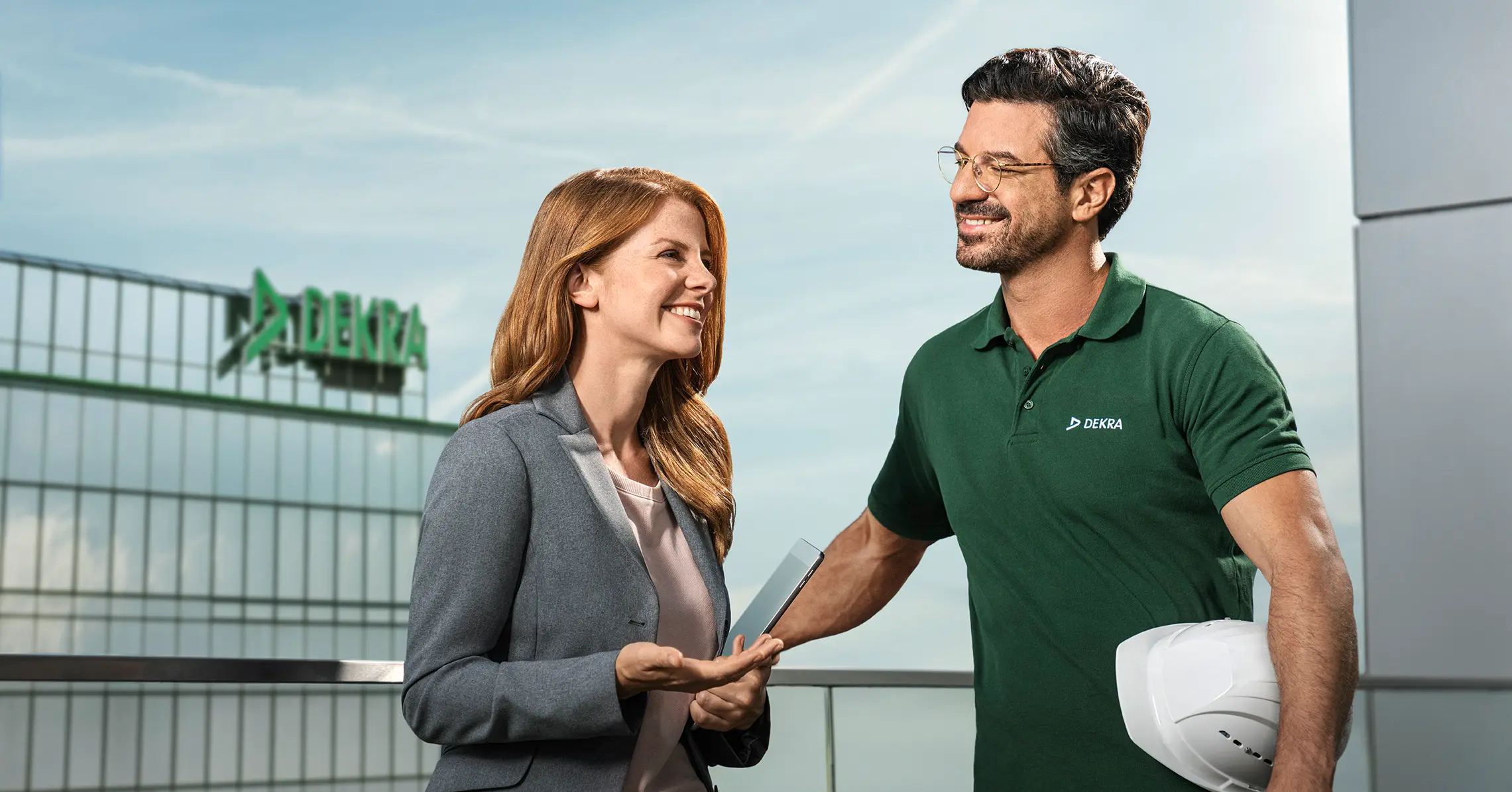 Mann im DEKRA Shirt und Frau im Business-Look unterhalten sich lächelnd vor einem DEKRA Gebäude