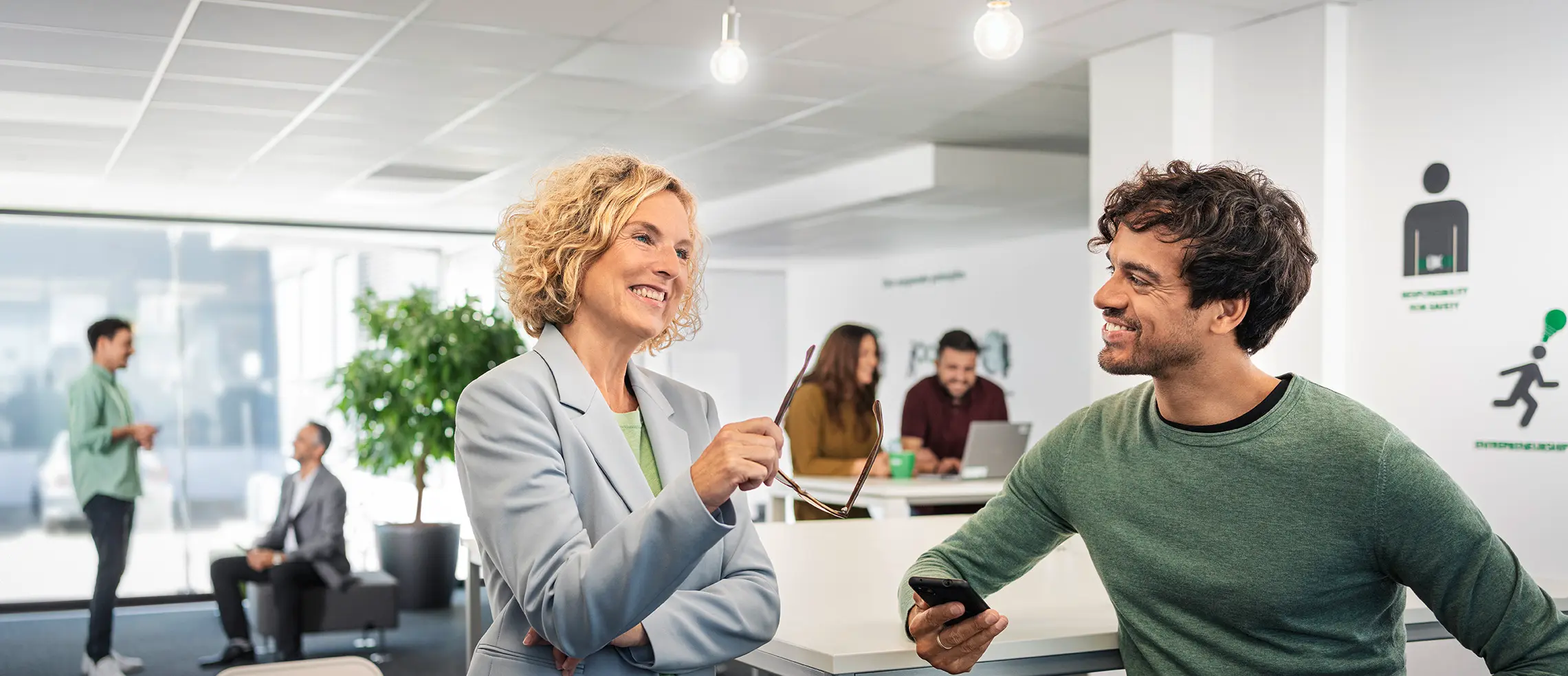 Zwei DEKRA Kollegen unterhalten sich in einem Open-Space-Bereich, weitere Mitarbeitende im Hintergrund