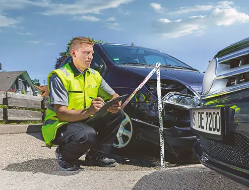 DEKRA Gutachter mit einem Zollstock neben ihm hockt vor zwei Unfallwagen und notiert etwas auf einem Klemmbrett