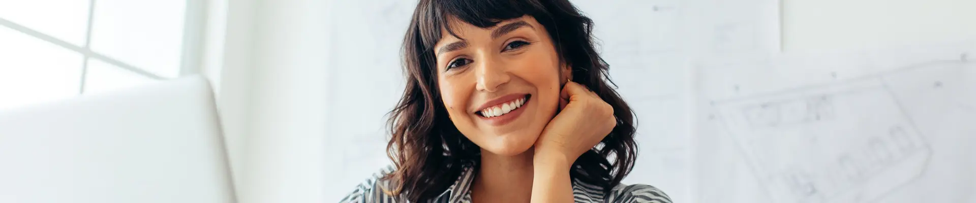 A woman smiles directly at the camera while sitting behind a laptop. Drawings and sketches are displayed on the wall behind her.