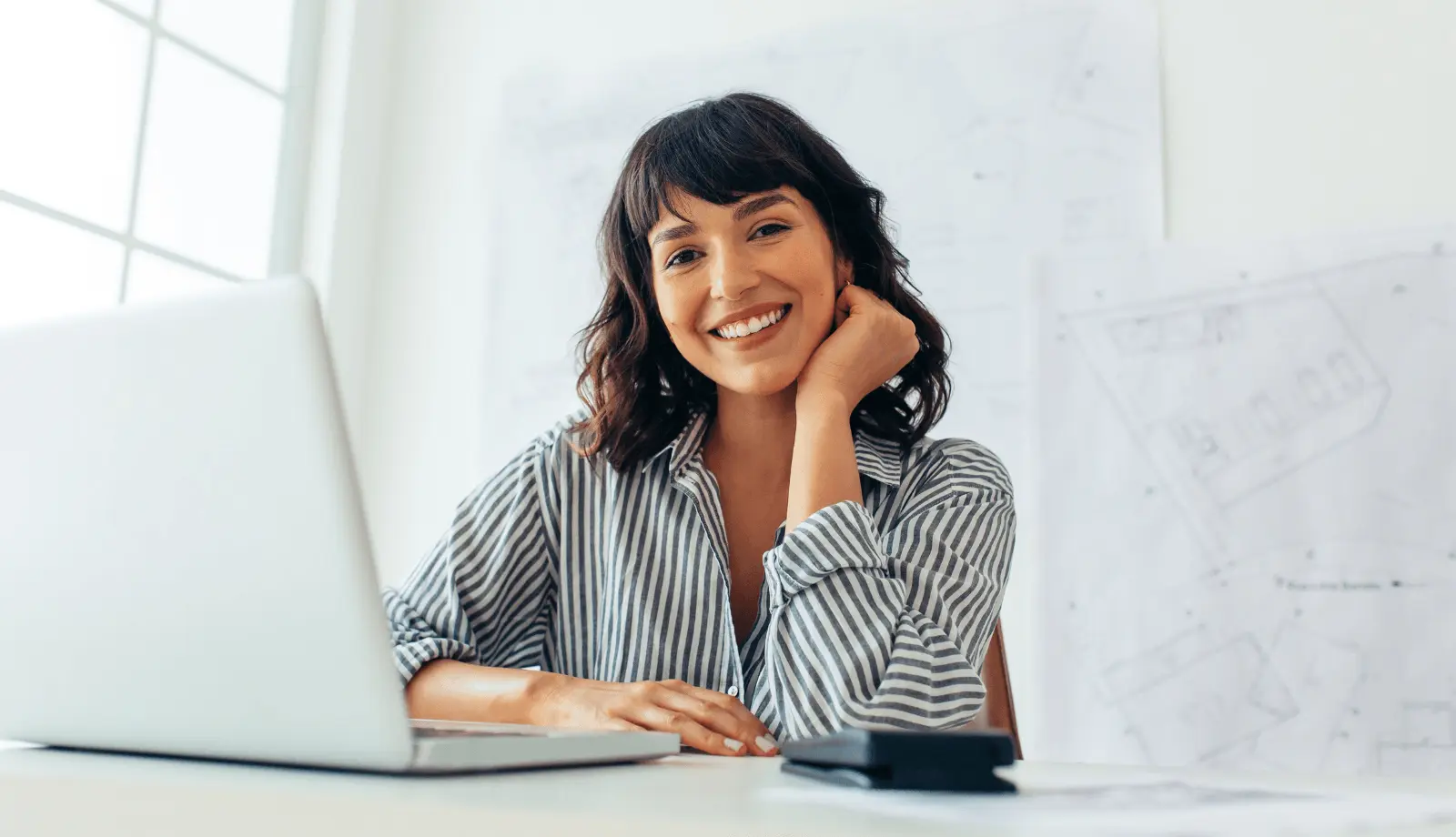 A woman smiles directly at the camera while sitting behind a laptop. Drawings and sketches are displayed on the wall behind her.