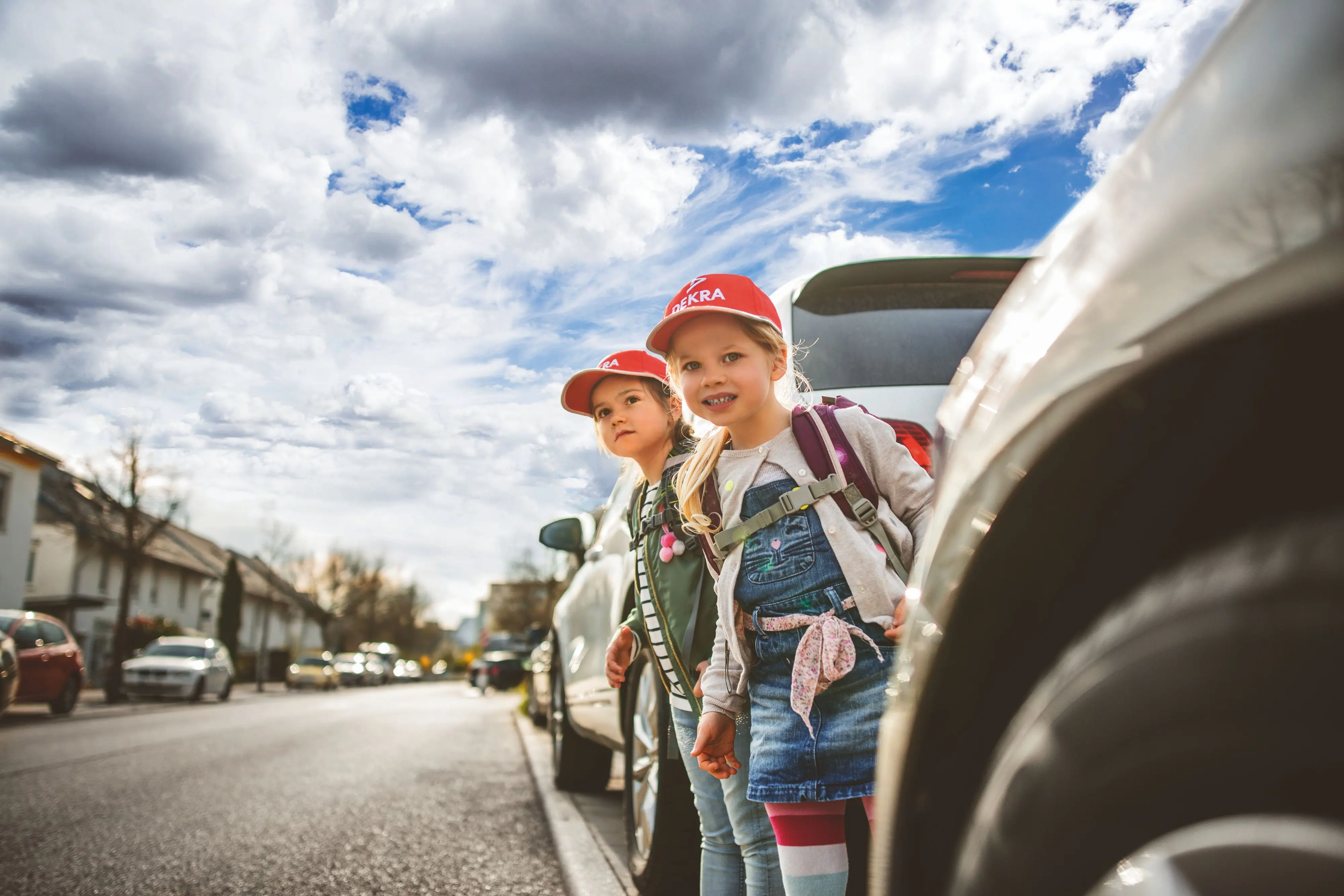 Zwei Kinder mit DEKRA Kinderkappen schauen zwischen zwei Autos über die Straße Zwei Kinder mit DEKRA Kinderkappen schauen zwischen zwei Autos über die Straße