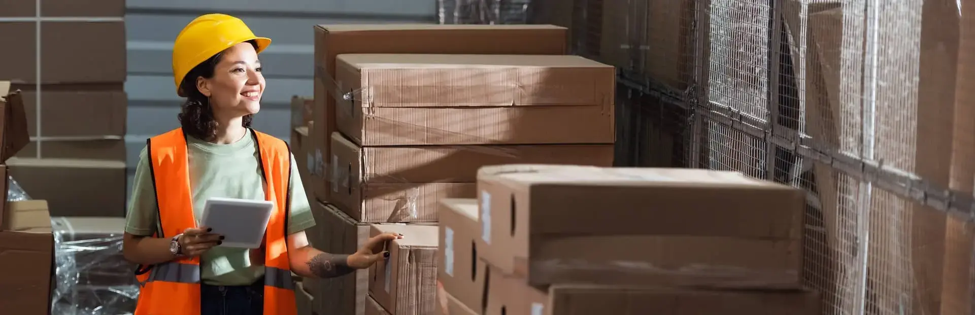  Pracownica magazynu w kamizelce i kasku kontrolująca etykiety przesyłek za pomocą tabletu – zgodność z wymaganiami IFS Logistics