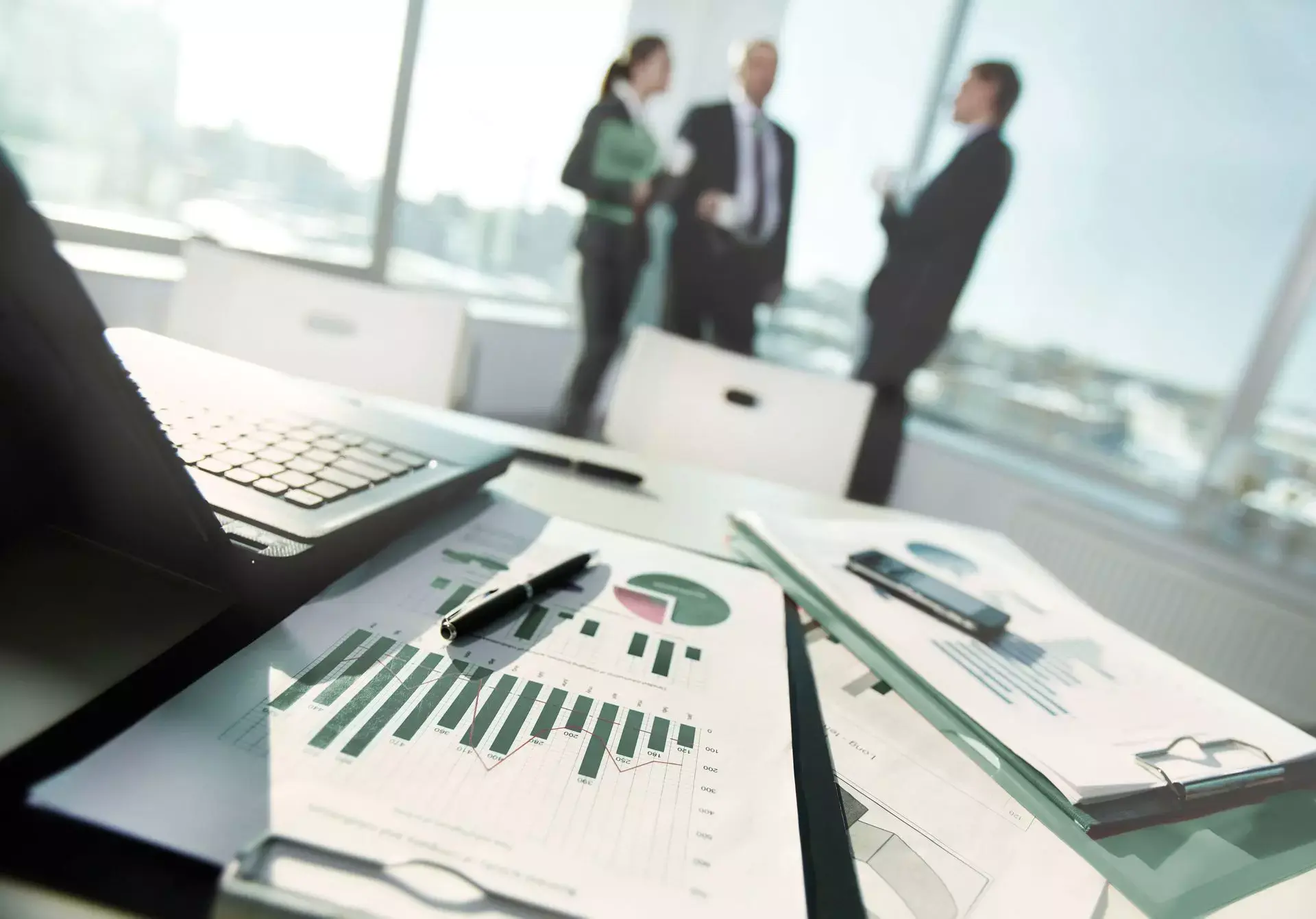 Dokumenty z wykresami na biurku, w tle rozmowa trzech osób w biurze, nawiązanie do weryfikacji i oceny dokumentacji technicznej.