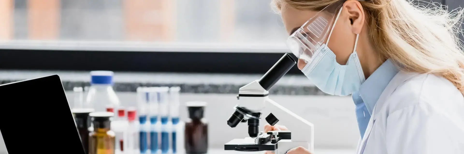 Naukowiec w masce medycznej patrzący przez mikroskop, symbolizujący higienę i bezpieczeństwo biologiczne