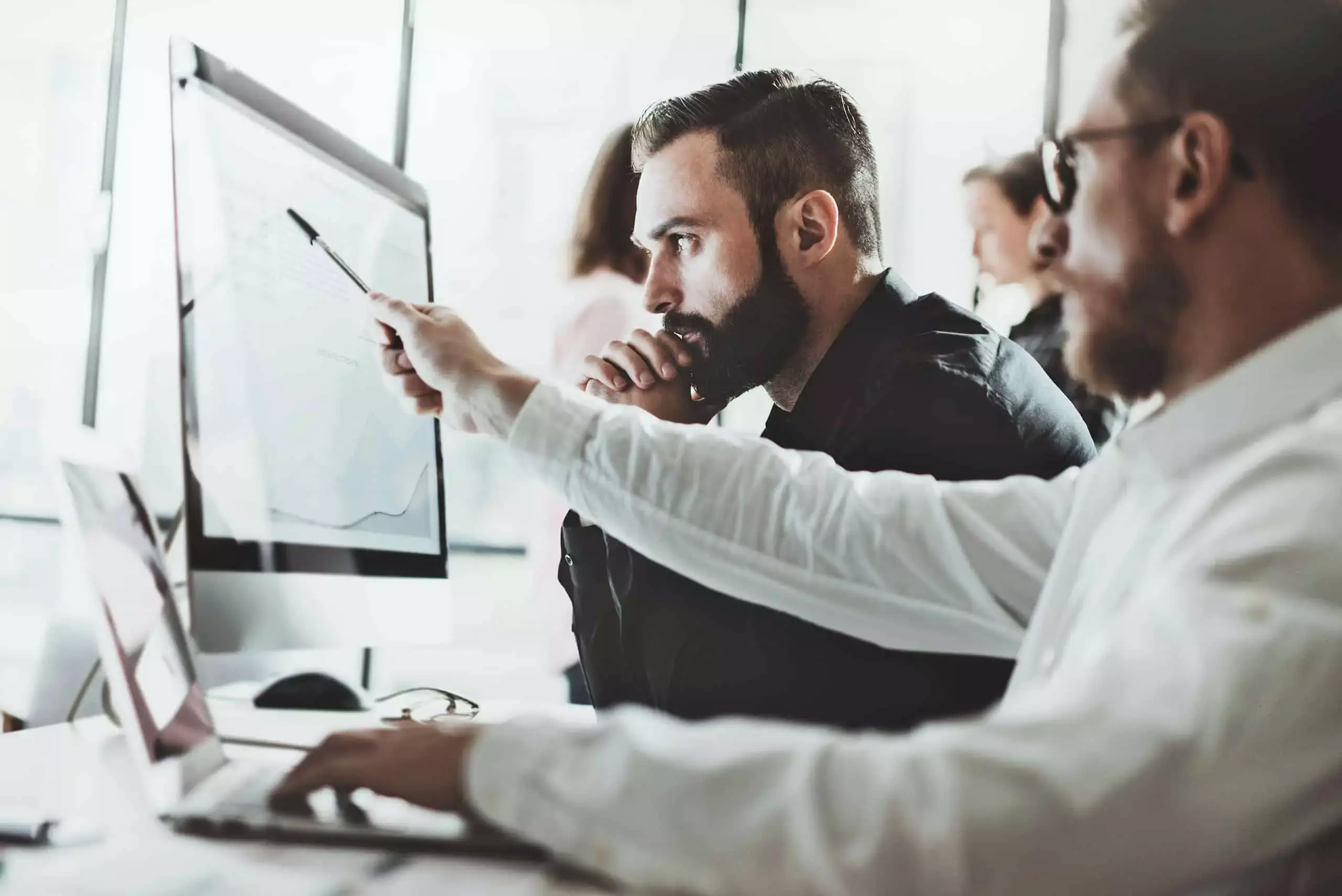 Dokumenty i wykresy na biurku, w tle osoby omawiające zarządzanie bezpieczeństwem informacji