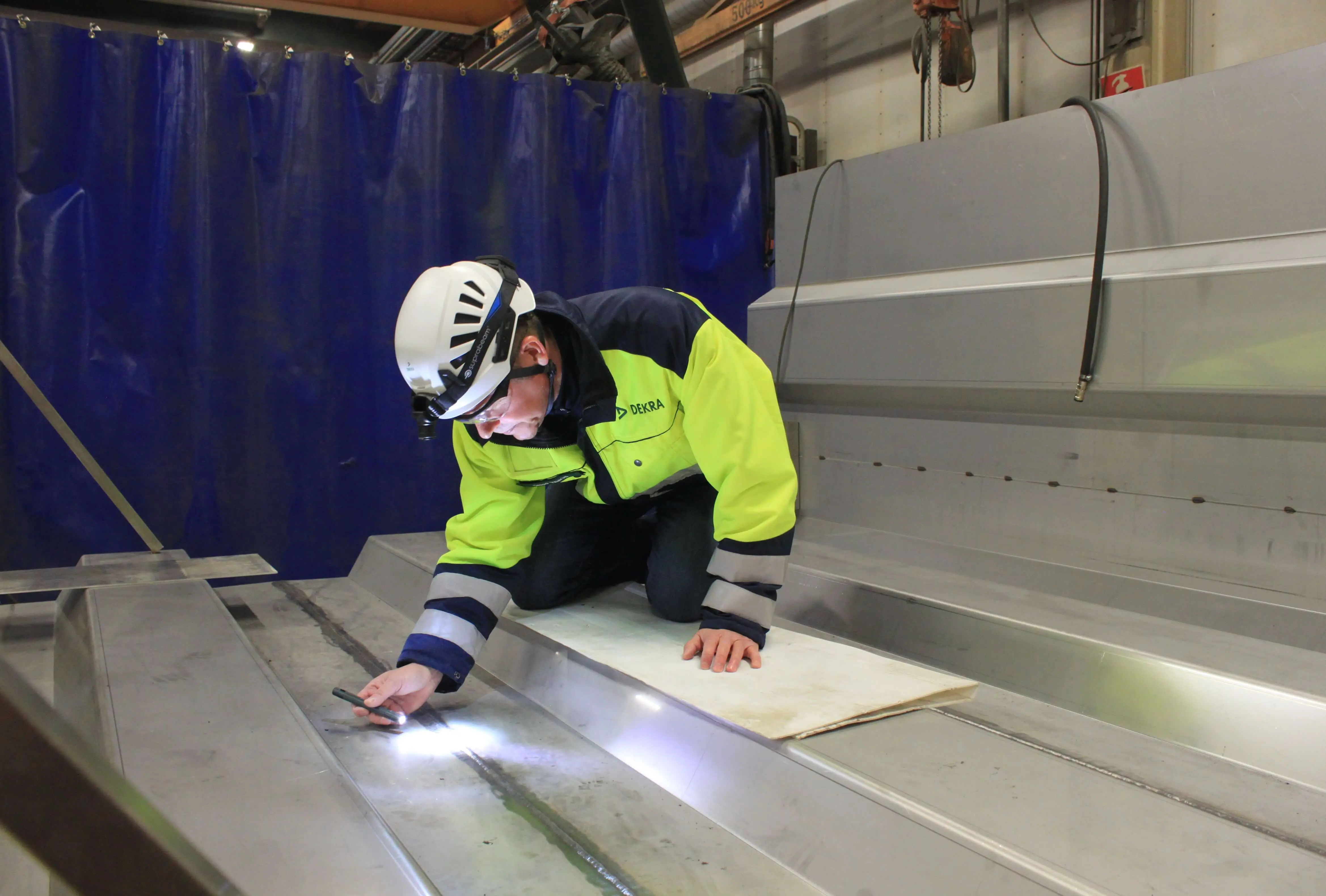 DEKRA-tarkastaja suorittamassa rikkomatonta testausta (NDT) metallikomponenteille, käyttäen taskulamppua pinnan vikojen havaitsemiseksi teollisessa ympäristössä.