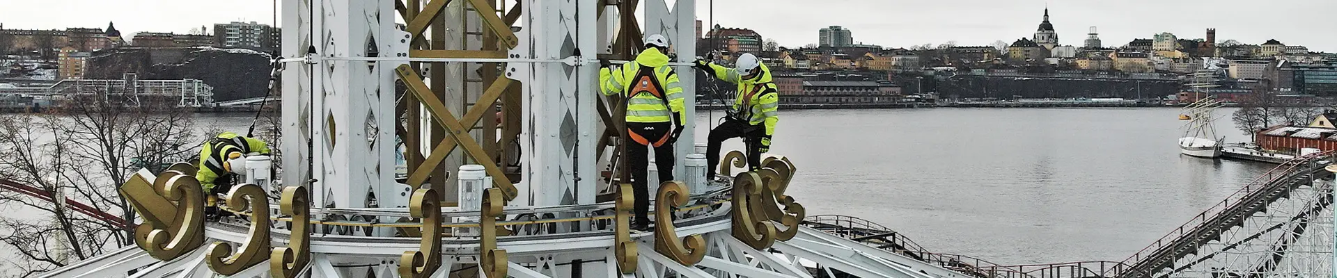  Två besiktningsingenjörer står högt upp i en åkatraktion på ett tivoli, med en livlig stadssilhuett synlig i bakgrunden.
