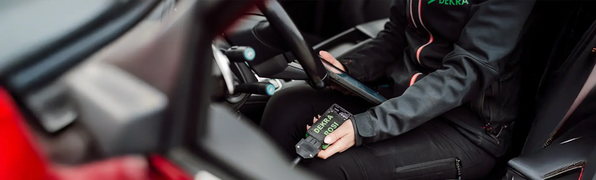 En svartklädd person som sitter i förarsätet i en bil och håller i en ipad och en dosa för batteritest.