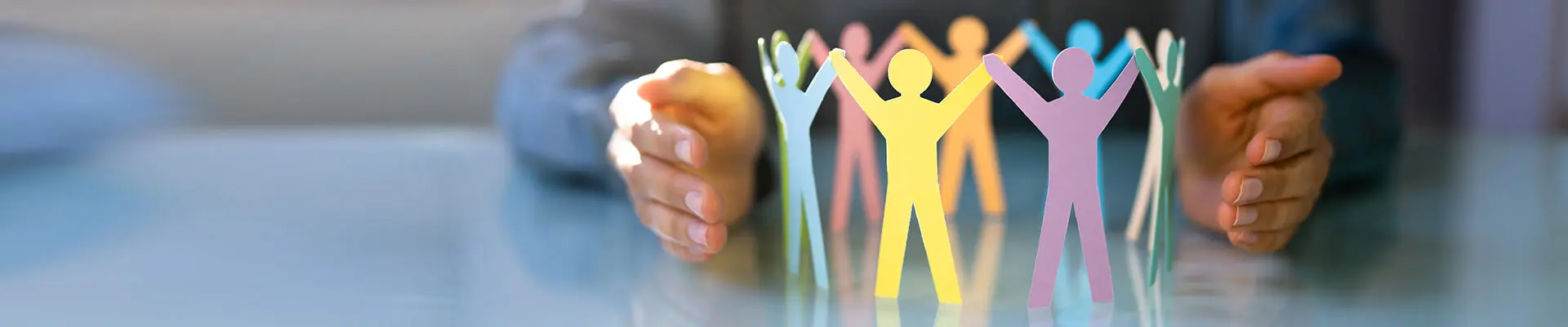 En person håller upp ett urklippt papper där små pappersfigurer, som står på rad och håller varandras händer, är utskurna. Pappersfigurerna ser ut som om de bildar en kedja av människor som håller varandra i händerna, symboliserande gemenskap och sammanhållning.