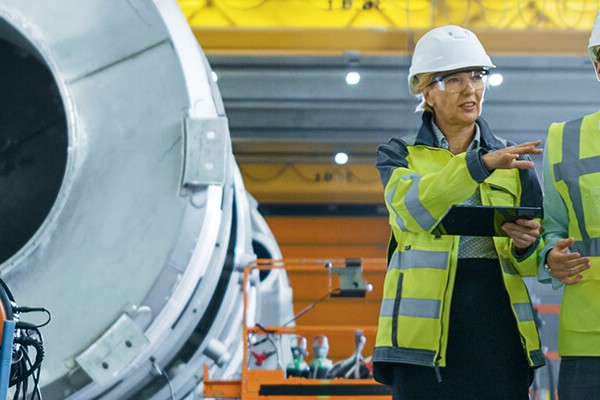 En man och en kvinna med reflexvästar går och kontrollerar byggnaden i en industriell miljö.