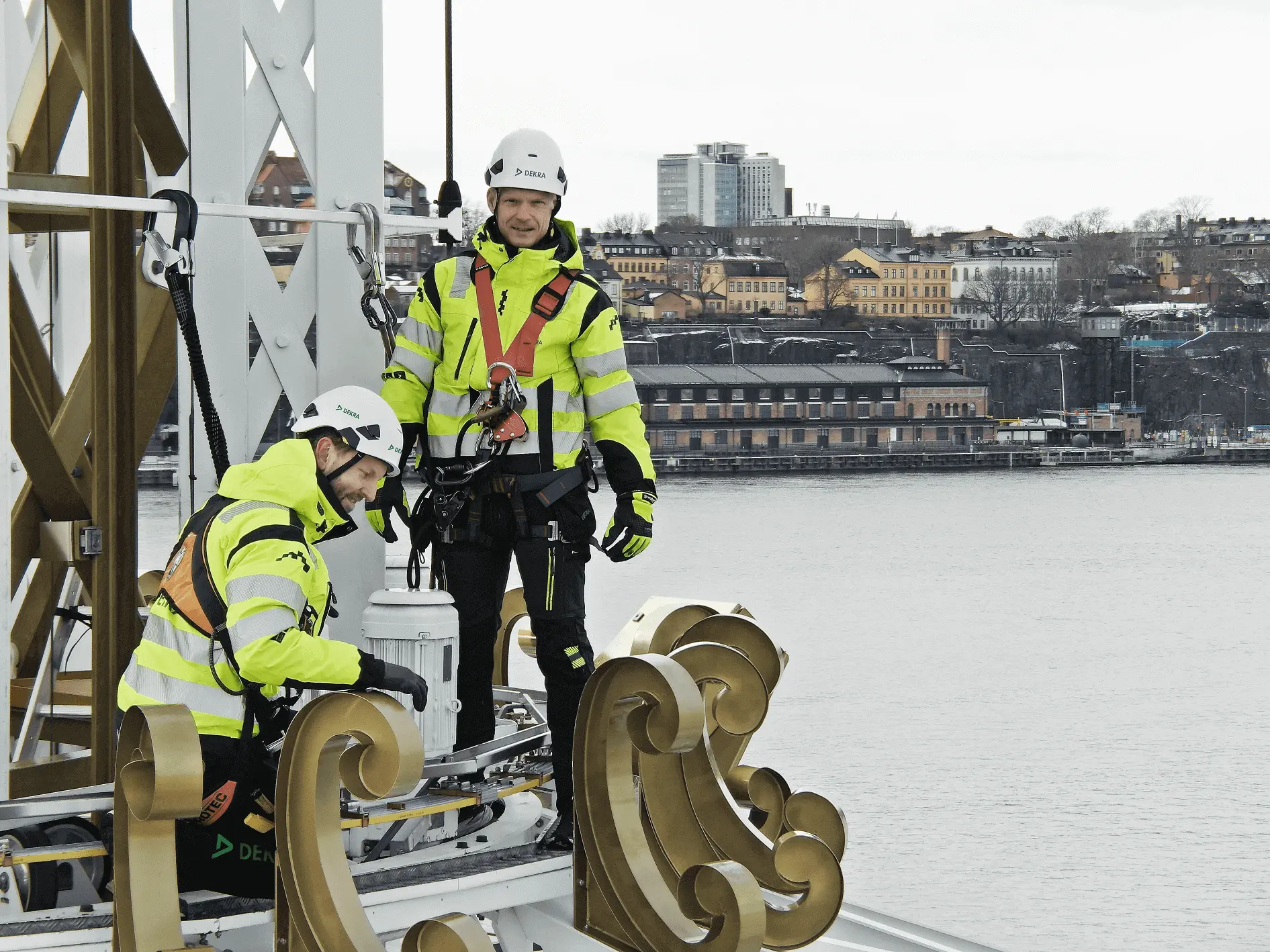 Två besiktningstekniker i gula reflexkläder och selar som står högt uppe i en åkatraktion med vatten och stad i bakgrunden. Två besiktningstekniker i gula reflexkläder och selar som står högt uppe i en åkatraktion med vatten och stad i bakgrunden.