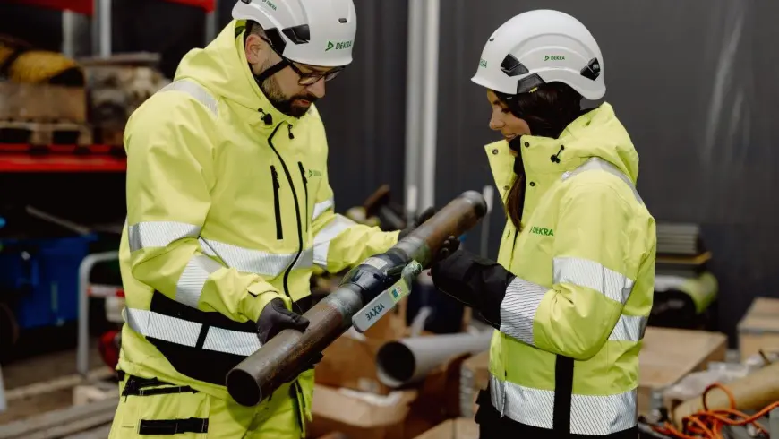 Två provningstekniker i gula varselkläder och vit hjälm som inspekterar ett rör.