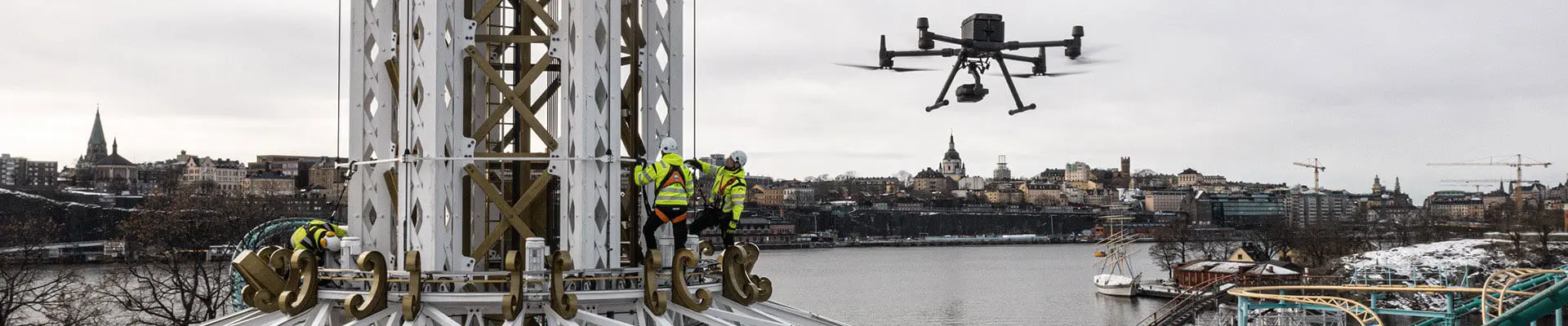 En drönare som flyger högt över ett nöjesfält med en attraktion, vatten och stad i bakgrunden.