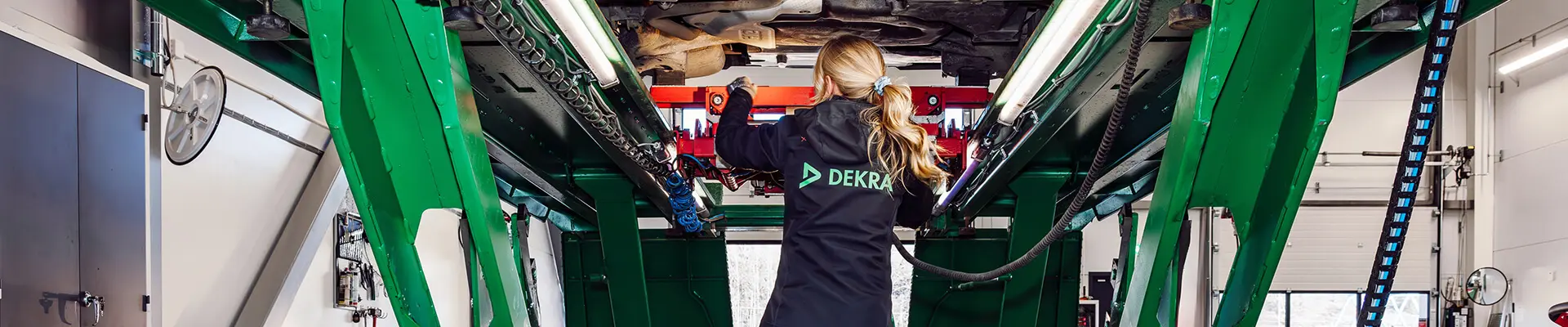 A woman in black work clothes stands under a car raised on a lift and conducts a car inspection.