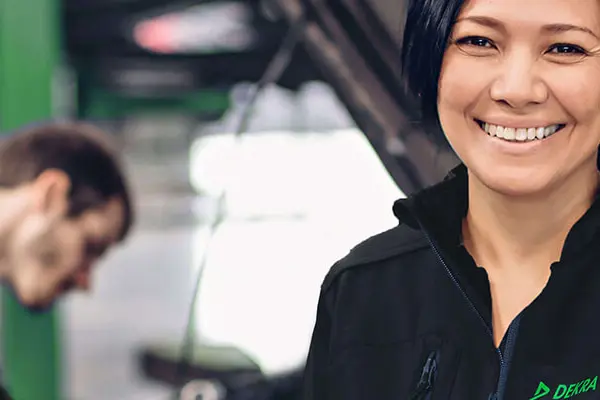 A woman in black work clothes stands in front of a background in the form of a car workshop with a man working on a picture engine with the hood open.