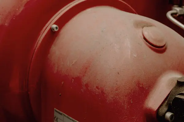 A dusty red boiler with a pressure gauge on it in an industrial premises.