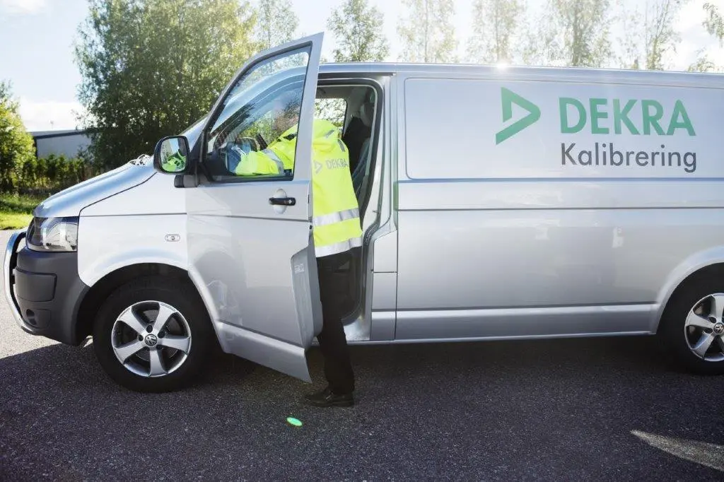 A man in reflective work clothes stands in the car door of a silver van that says DEKRA Kalibring..