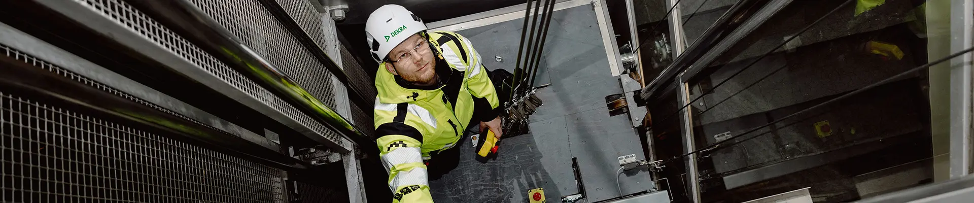  En man i en gul jacka står på taket av en hiss inne i ett hisschakt och tittar uppåt i hisschaktet.