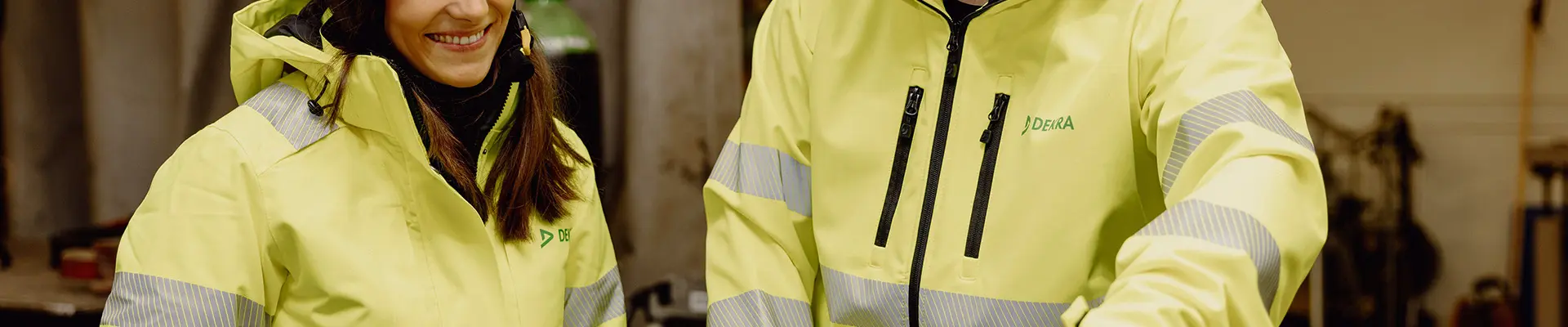 Två personer i reflekterande arbetskläder står vid en laptop och pratar i en verkstadslokal. Två personer i reflekterande arbetskläder står vid en laptop och pratar i en verkstadslokal.