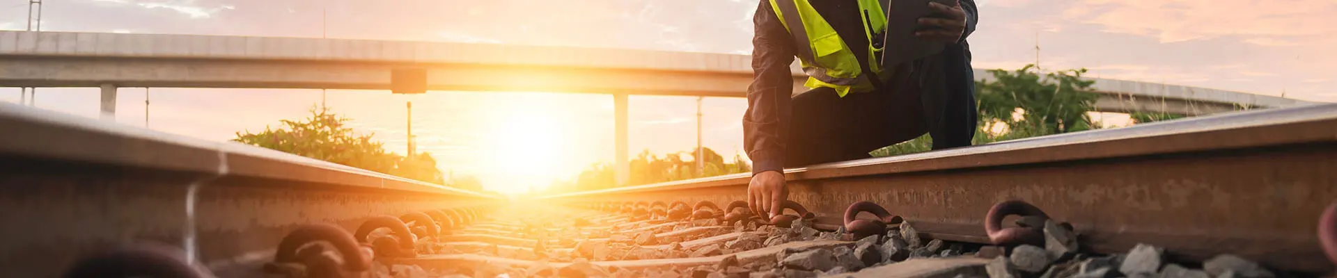 En man i reflekterande arbetskläder sitter på huk vid ett tågspår och inspekterar rälsen i solnedgång.