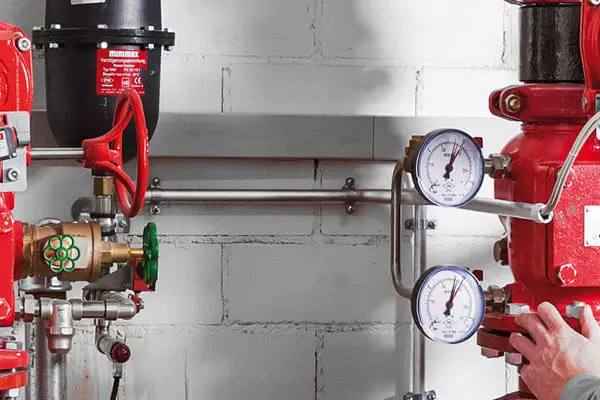 A man in work clothes stands and inspects several red pipes with compressed air and pressure gauges on them.