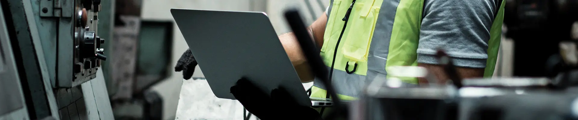 An industrial worker in a safety vest uses a laptop near a machine, highlighting the integration of technology in industrial work.