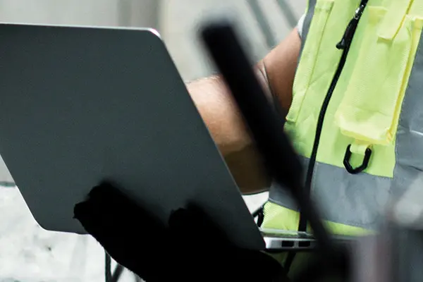 An industrial worker in a safety vest uses a laptop near a machine, highlighting the integration of technology in industrial work.